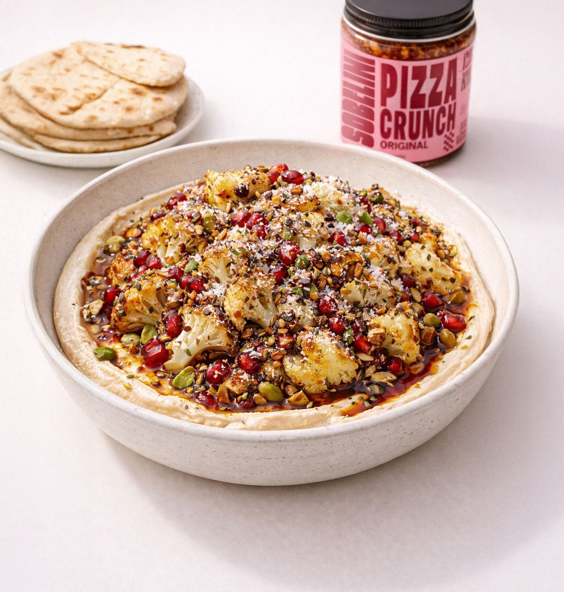 Roasted cauliflower florets layered over creamy hummus, topped with pistachios, pomegranate seeds, za’atar, and a glossy drizzle of pomegranate molasses and Sobelini Pizza Crunch, served in a shallow bowl with warm pita on a neutral backdrop.