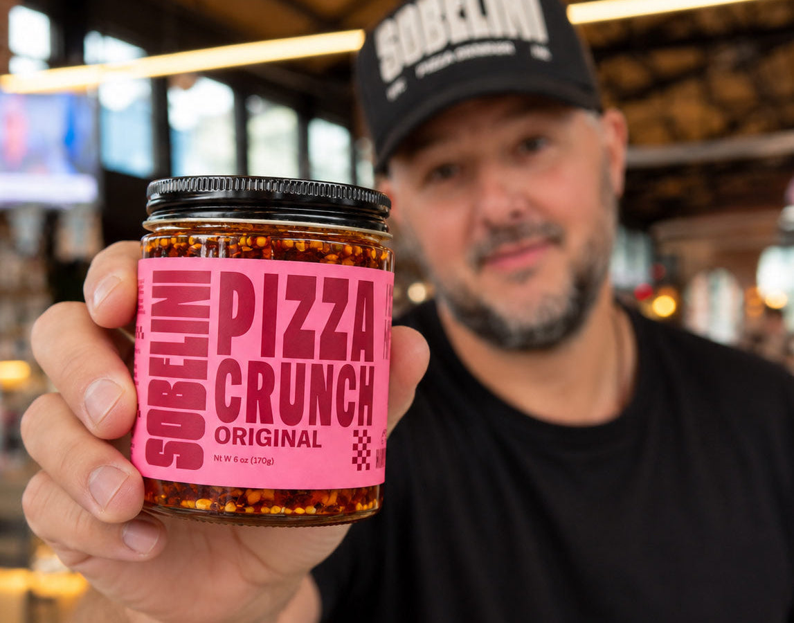 A man wearing a black cap with 'SOBELINI' on it holds up a jar of Sobelini Pizza Crunch Original to the camera in what appears to be an indoor market or restaurant setting.